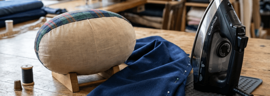 Tailoring tools on wooden table including fabric, iron, pins, thimble, and measuring tape